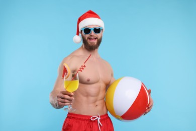 Photo of Attractive young man with muscular body in Santa hat holding ball and cocktail on light blue background