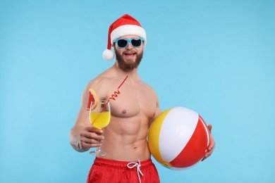 Attractive young man with muscular body in Santa hat holding ball and cocktail on light blue background