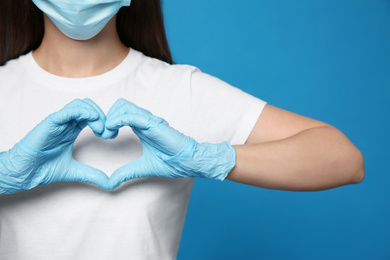 Photo of Female volunteer in protective mask and gloves showing heart gesture on blue background, closeup. Aid during coronavirus quarantine
