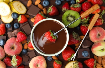 Photo of Fondue fork with strawberry in bowl of melted chocolate surrounded by other fruits on wooden table, flat lay