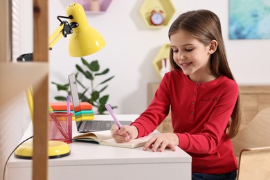 Photo of E-learning. Cute girl taking notes during online lesson at table indoors