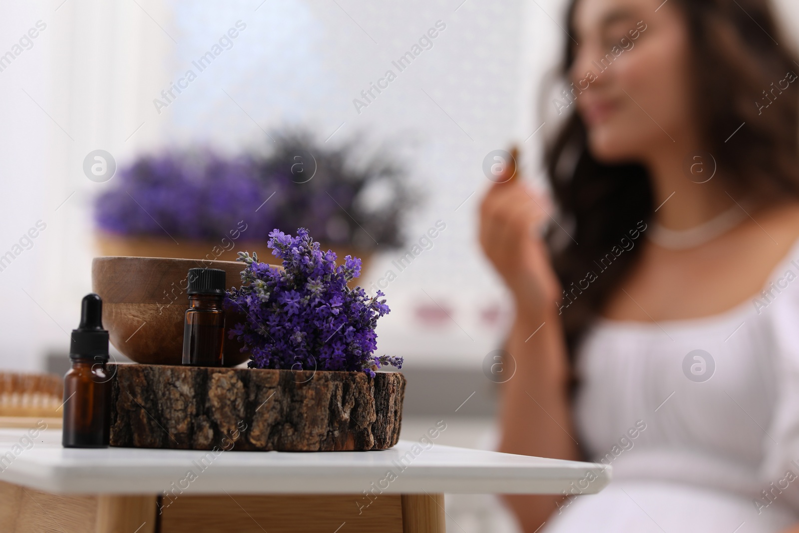 Photo of Woman sitting at table indoors, focus on stump with bottles of essential oil and flowers