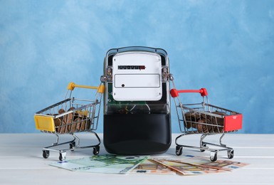 Electricity meter, small shopping carts with coins and euro banknotes on white wooden table