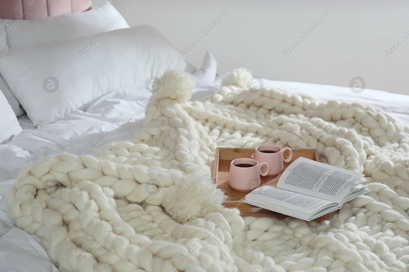 Photo of Wooden tray with cups and book on white knitted plaid in bedroom