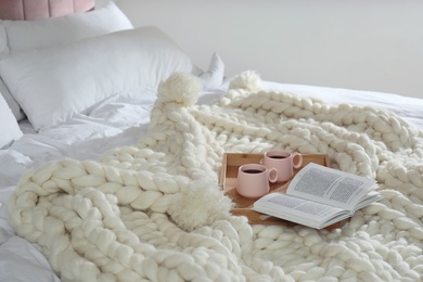 Wooden tray with cups and book on white knitted plaid in bedroom