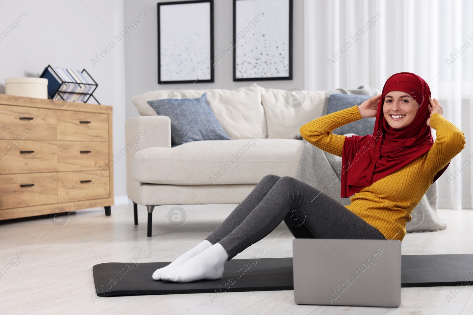 Photo of Muslim woman in hijab doing abs exercise near laptop on fitness mat at home. Space for text