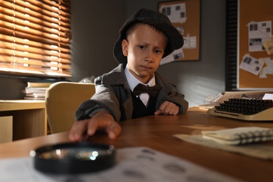Cute little detective taking magnifying glass at table in office