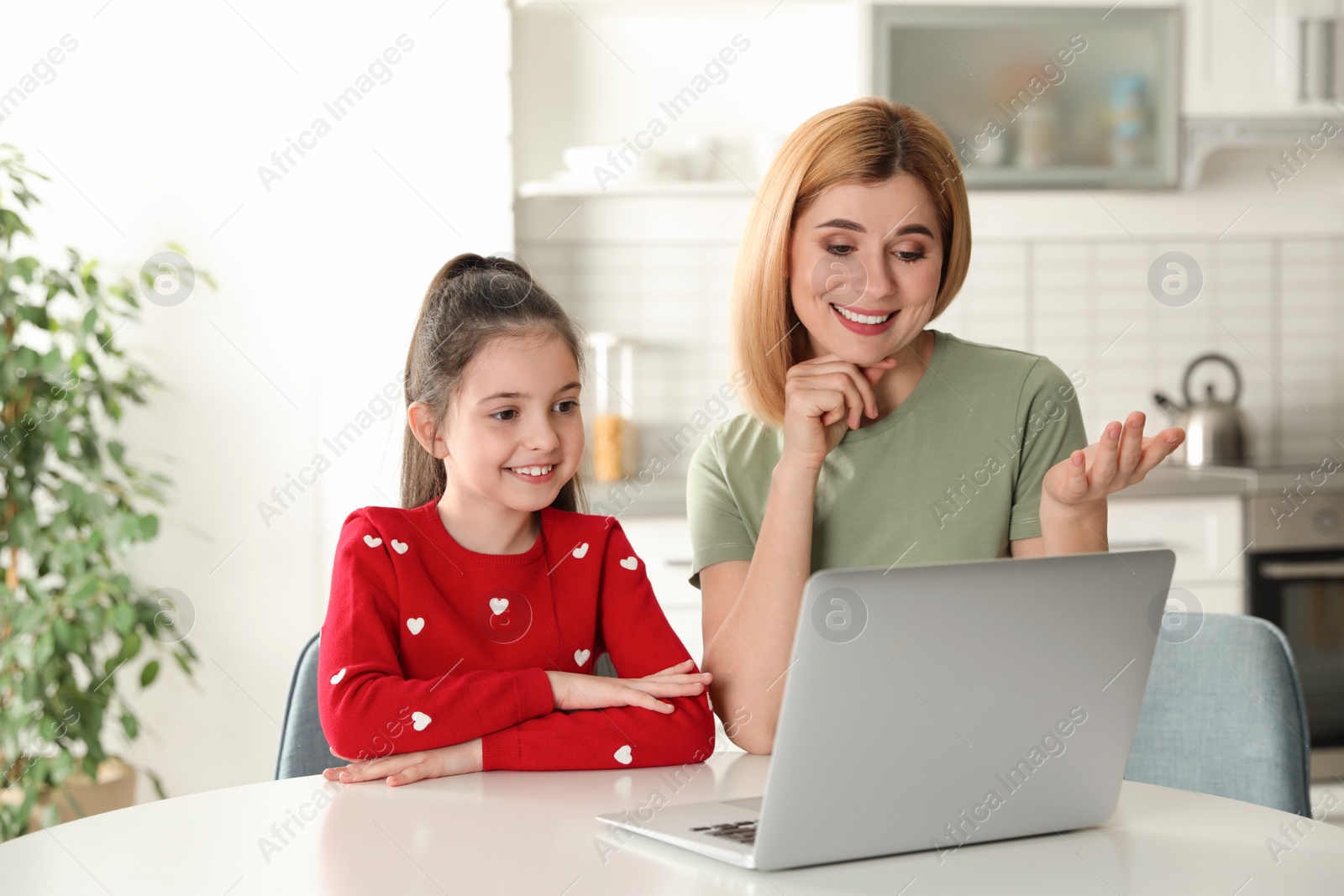 Photo of Mother and her daughter using video chat on laptop at table in kitchen