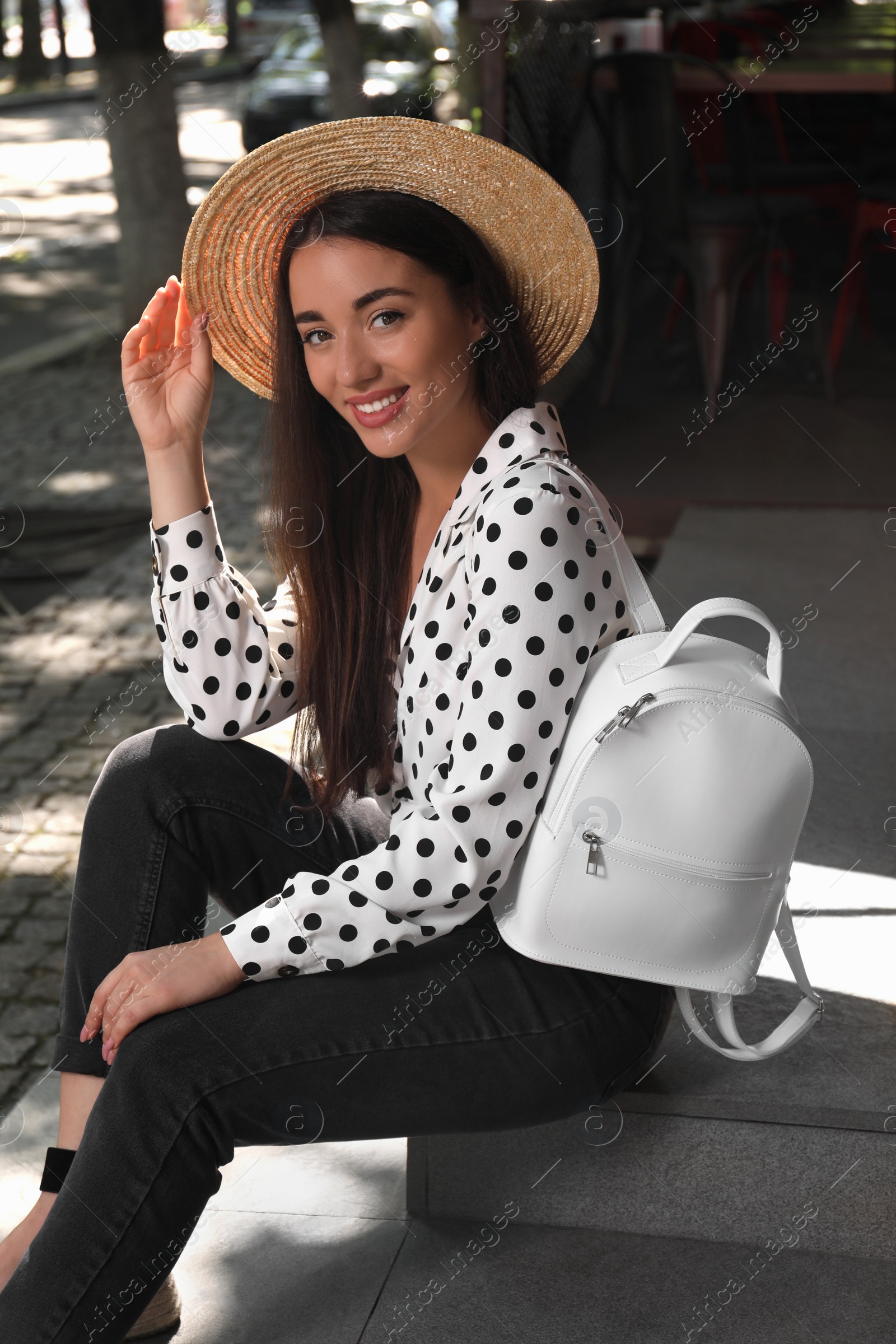 Photo of Beautiful young woman with stylish backpack and hat sitting on stairs outdoors