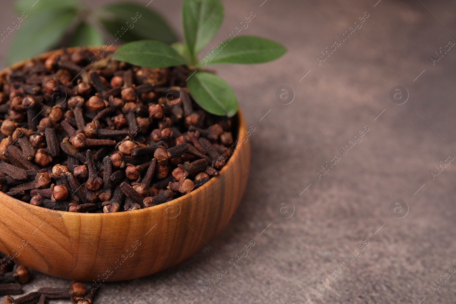 Photo of Aromatic cloves and green leaves in bowl on brown table, closeup. Space for text