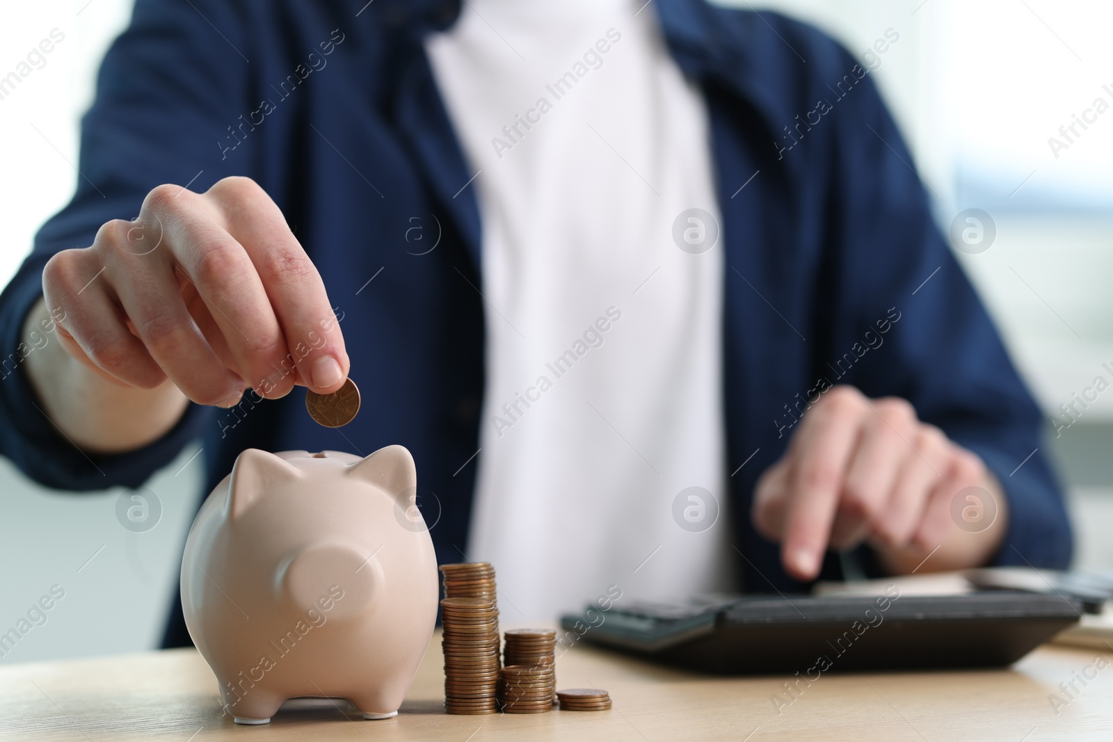 Photo of Financial savings. Man putting coin into piggy bank while using calculator at wooden table, closeup