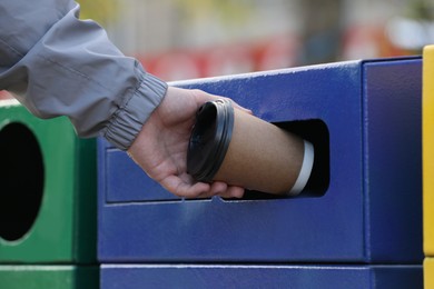 Photo of Man throwing paper coffee cup into garbage bin outdoors, closeup. Waste sorting