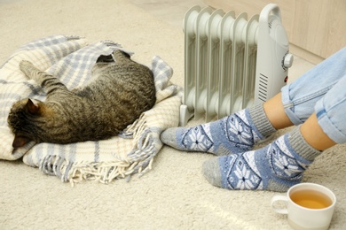 Photo of Young woman and cute tabby cat near electric heater at home, closeup