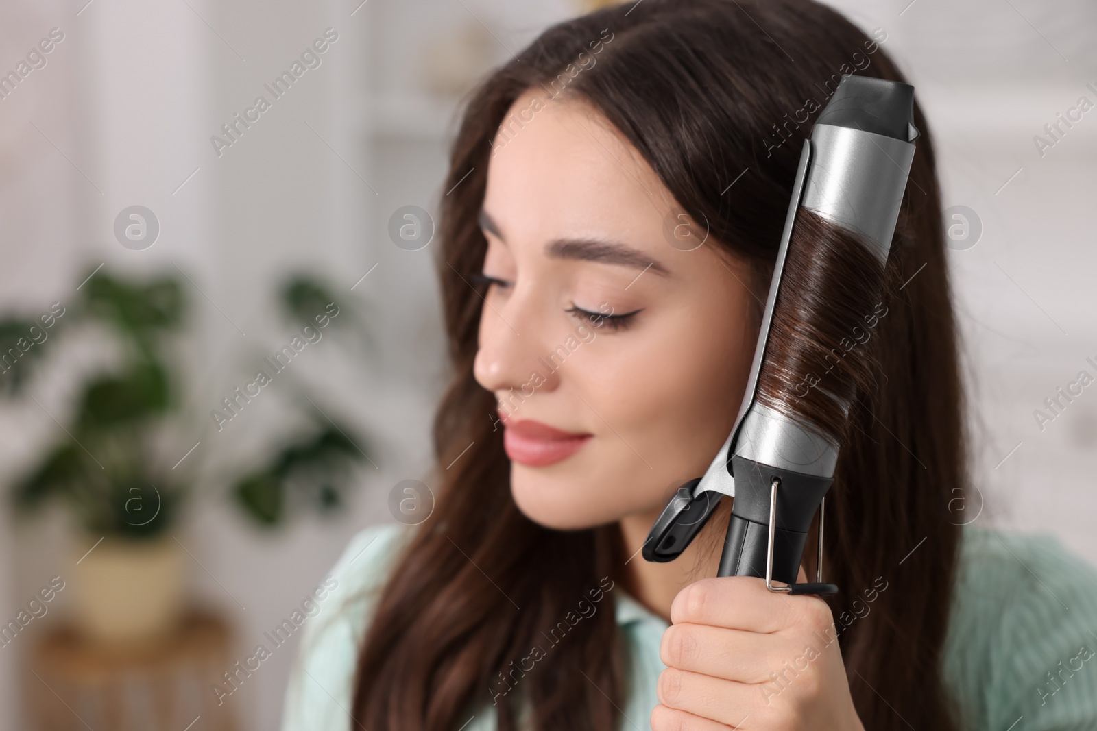 Photo of Beautiful woman using curling hair iron indoors. Space for text