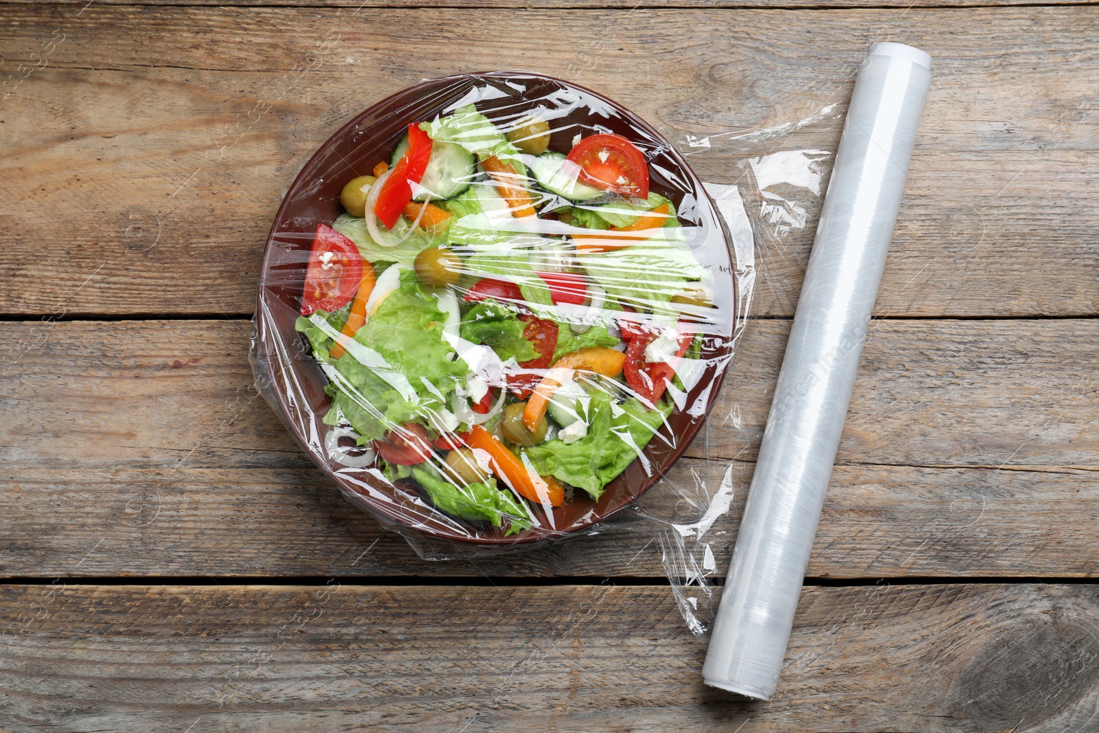 Photo of Bowl of fresh salad with plastic food wrap on wooden table, flat lay