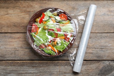 Photo of Bowl of fresh salad with plastic food wrap on wooden table, flat lay