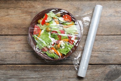 Bowl of fresh salad with plastic food wrap on wooden table, flat lay