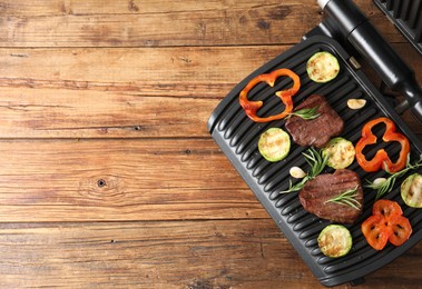 Photo of Electric grill with tasty meat steaks, rosemary and vegetables on wooden table, top view. Space for text