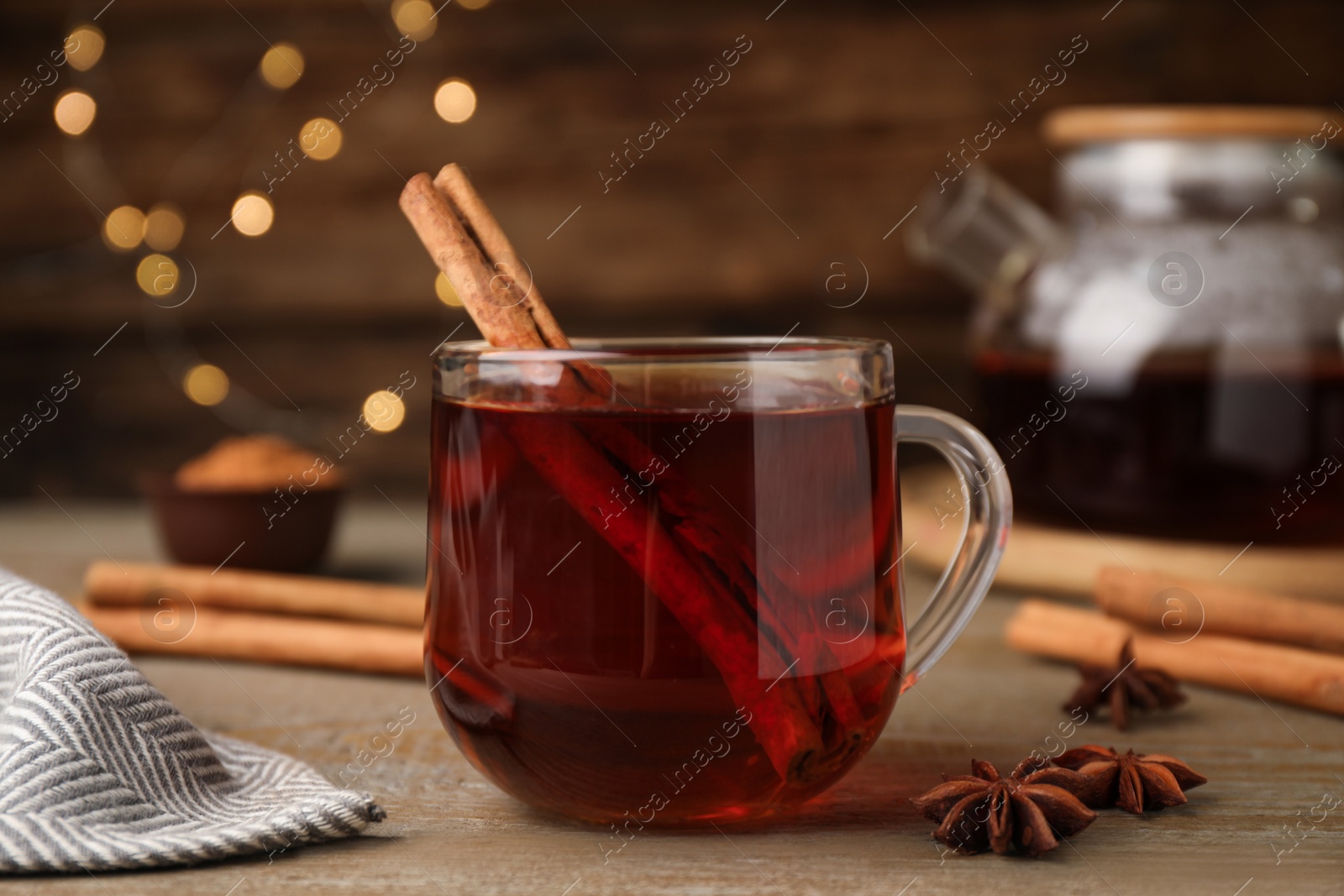 Photo of Glass cup of hot tea with aromatic cinnamon on wooden table against blurred lights