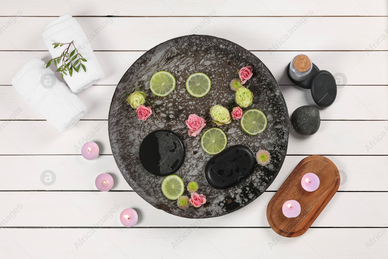 Photo of Plate with water, flowers, lime slices and burning candles on white wooden floor, flat lay. Pedicure procedure