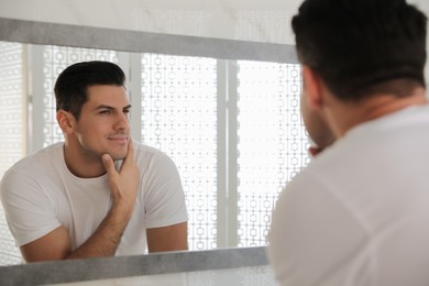 Photo of Handsome man touching his smooth face after shaving near mirror in bathroom