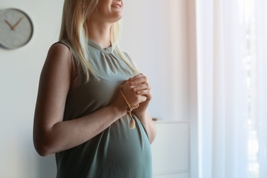 Young pregnant woman with beads praying at home. Space for text