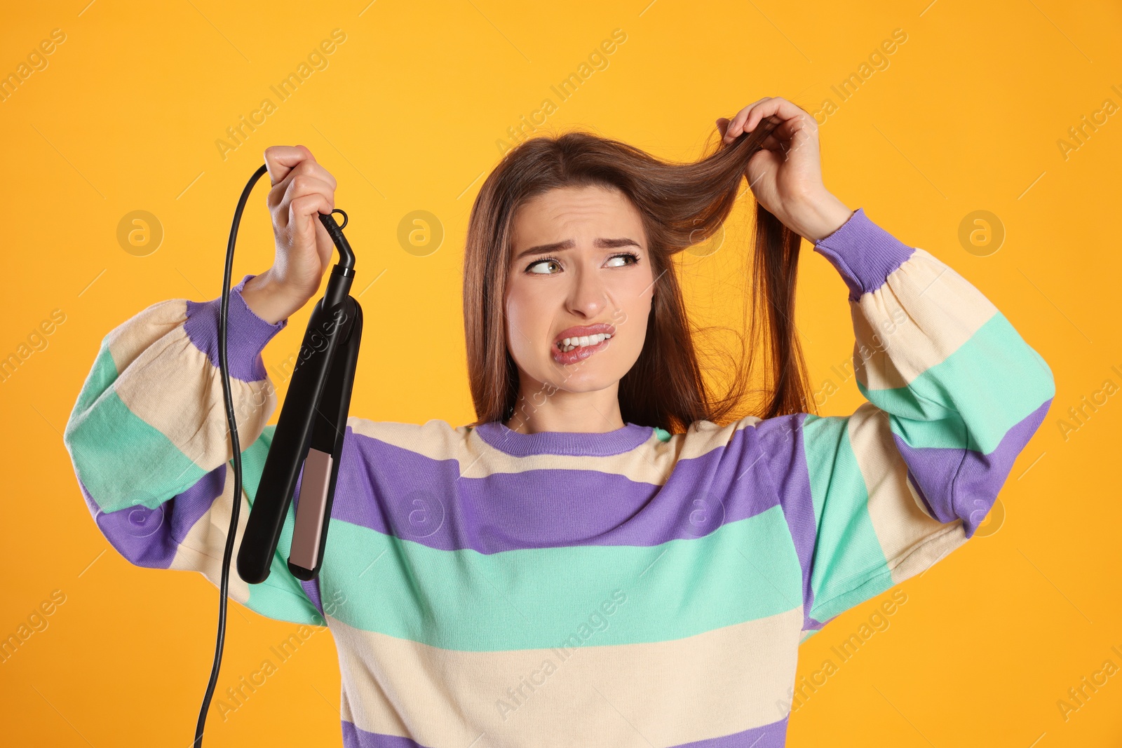 Photo of Stressed young woman with flattening iron on yellow background. Hair damage