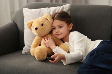 Little girl with toy bear changing TV channels by remote control on sofa at home