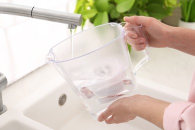 Photo of Woman filling filter jug with water from tap in kitchen, closeup