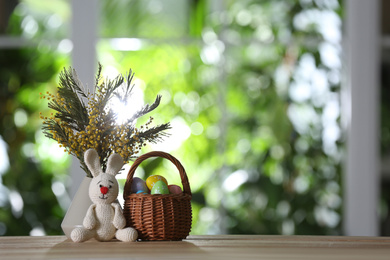 Photo of Easter bunny toy, dyed eggs and flowers on wooden table against blurred green background. Space for text