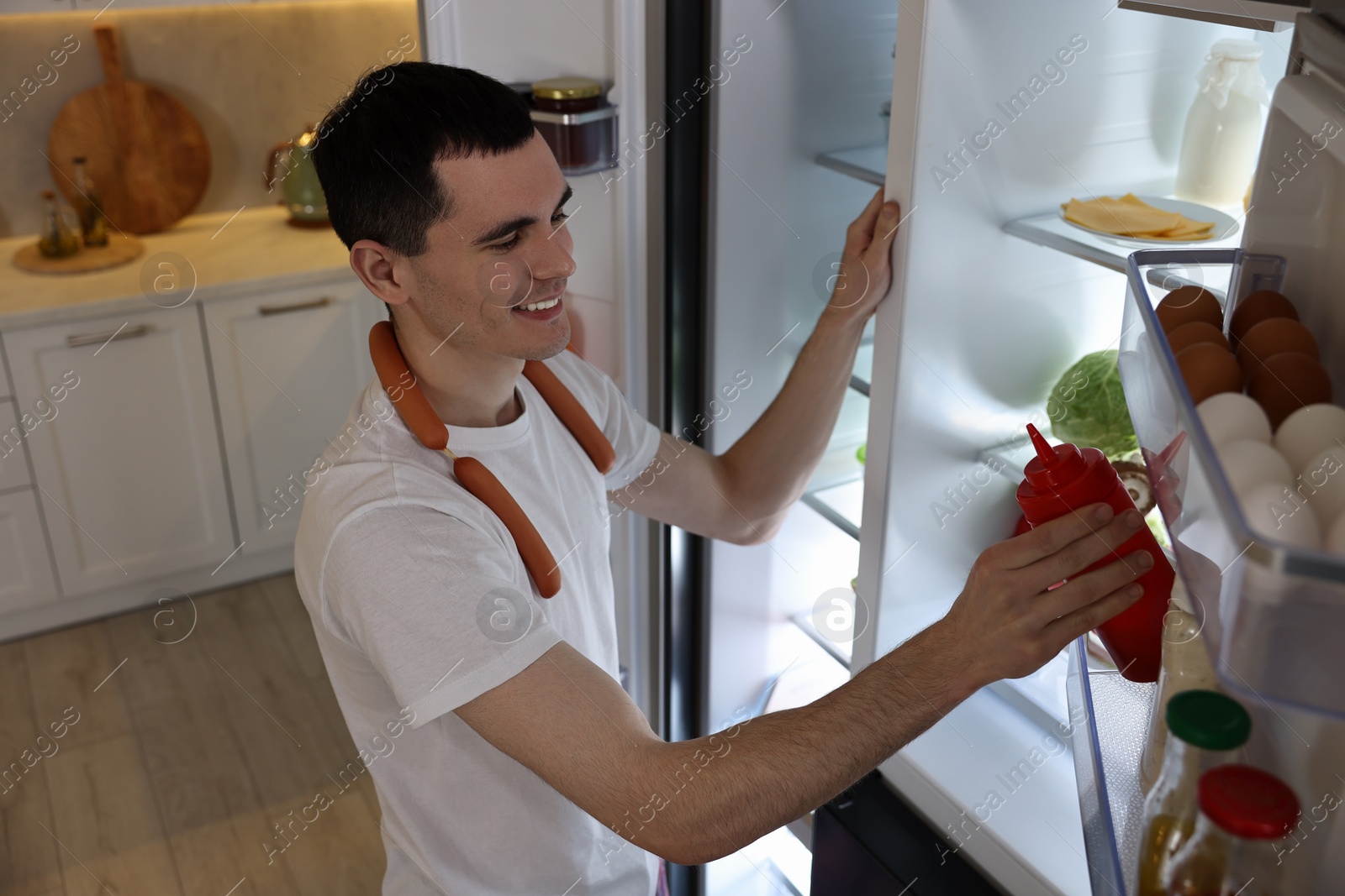 Photo of Happy man with sausages taking ketchup out of refrigerator in kitchen