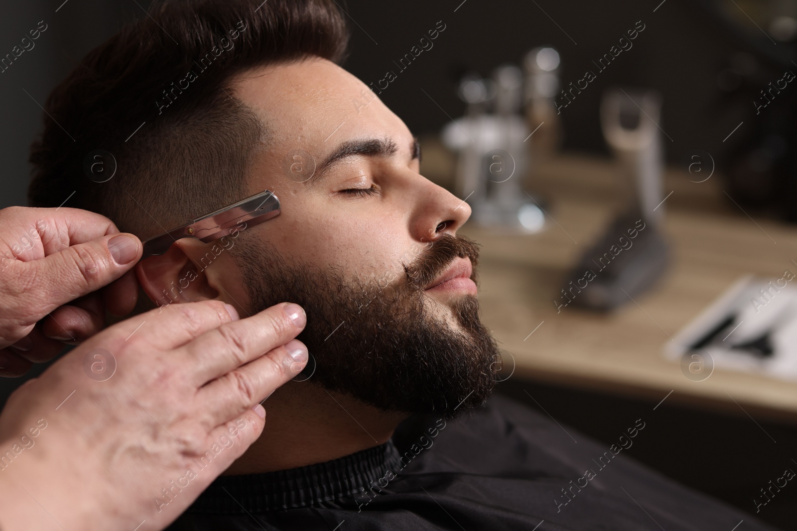 Photo of Professional barber shaving client's beard with blade in barbershop, closeup