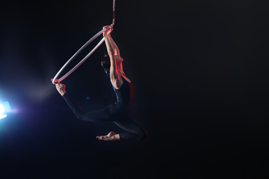 Young woman performing acrobatic element on aerial ring against dark background. Space for text