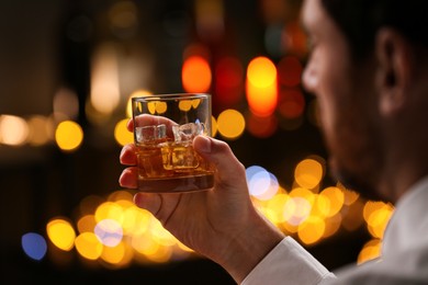 Man holding glass of whiskey with ice cubes against blurred lights, selective focus