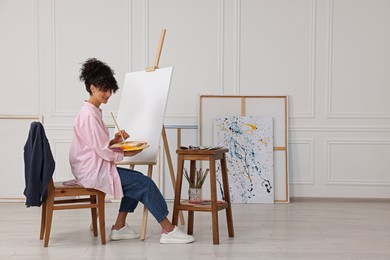 Photo of Young woman mixing paints on palette with brush near easel in studio. Space for text
