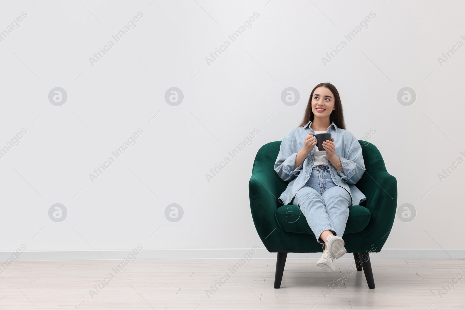 Photo of Beautiful woman with cup of drink sitting in armchair near white wall indoors, space for text