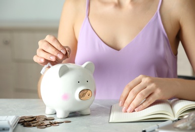 Photo of Woman putting coin into piggy bank at grey marble table, closeup. Money savings