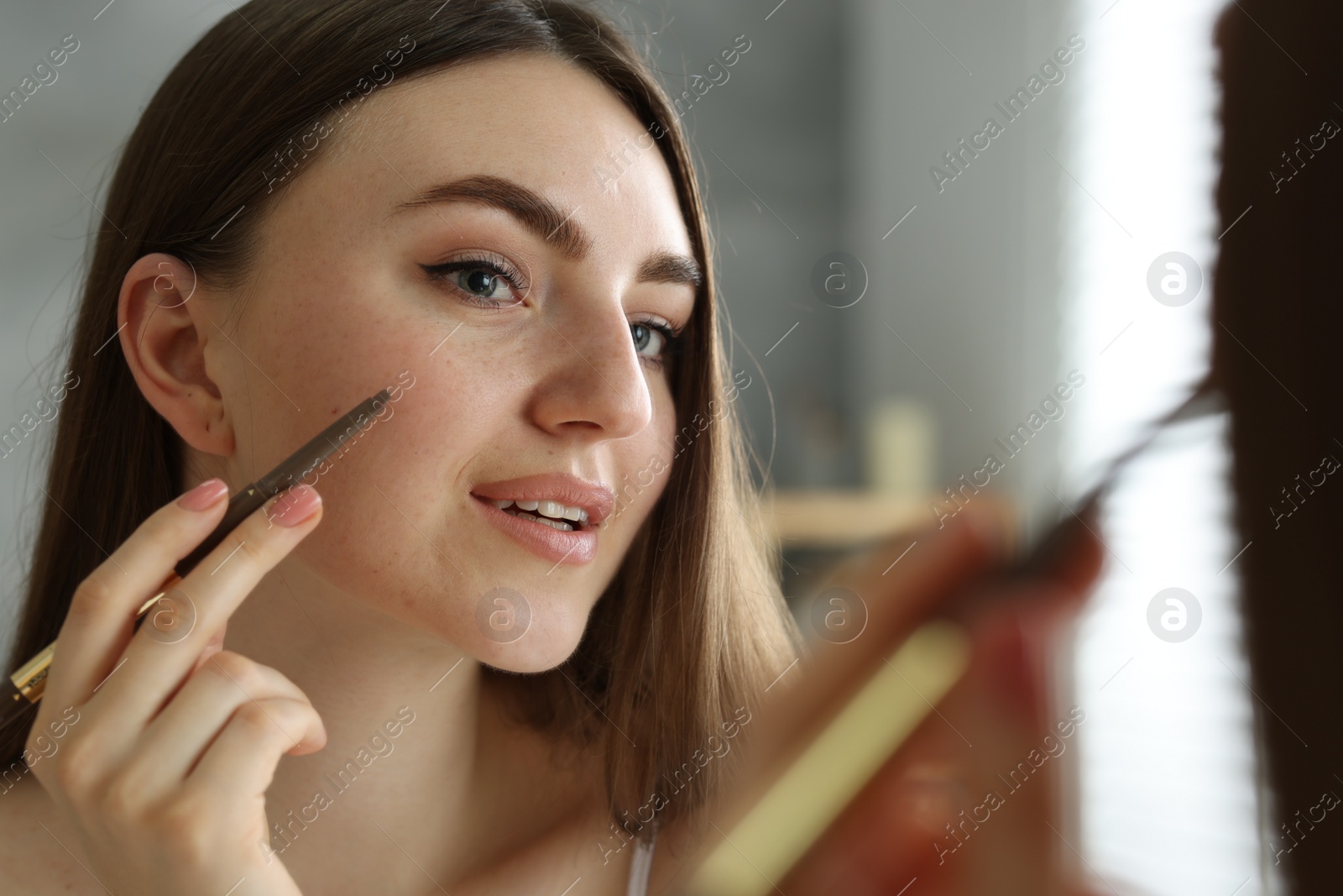 Photo of Beautiful woman drawing freckles with pen near mirror indoors, closeup