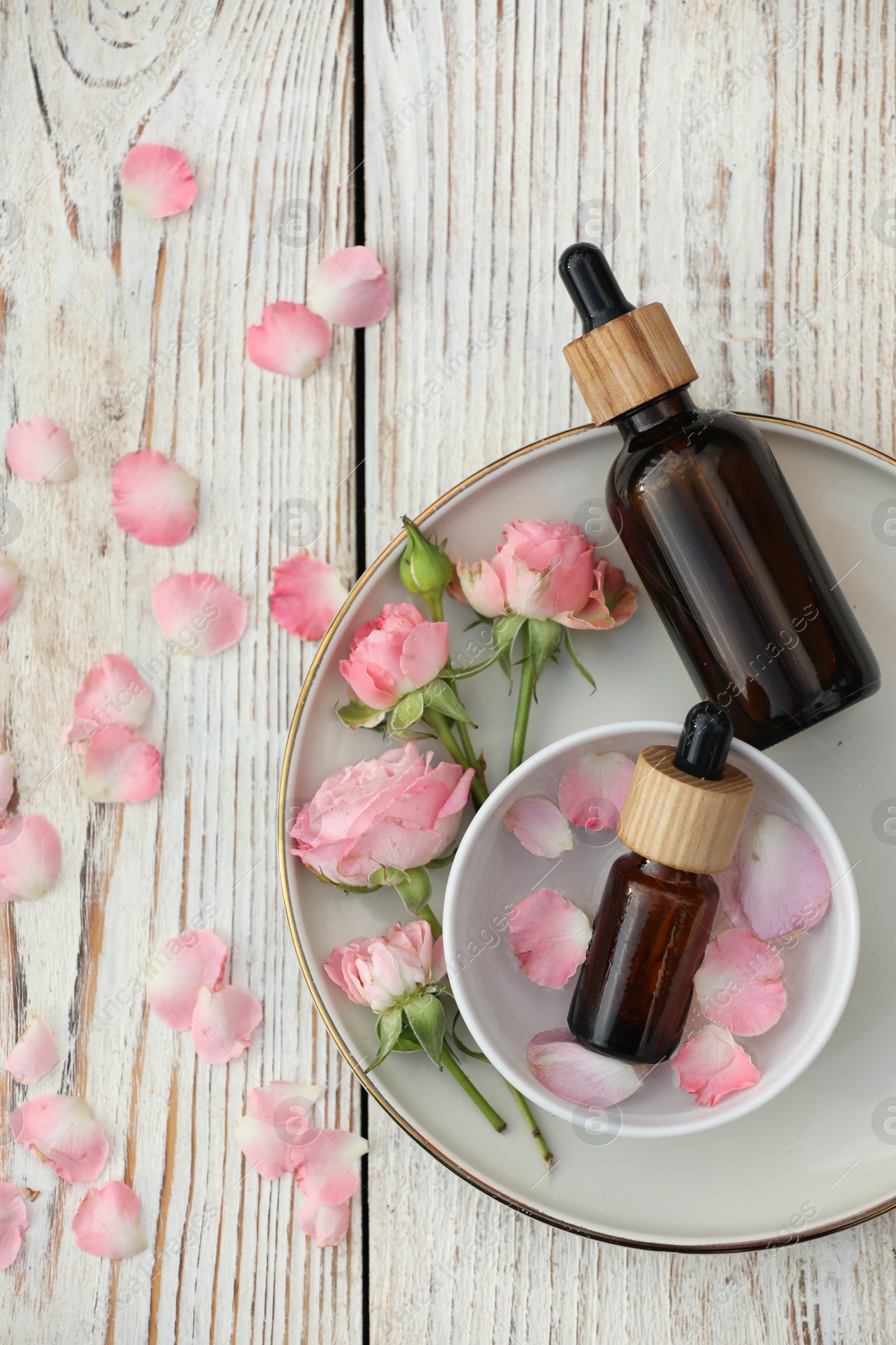 Photo of Bottles of rose essential oil and flowers on white wooden table, flat lay