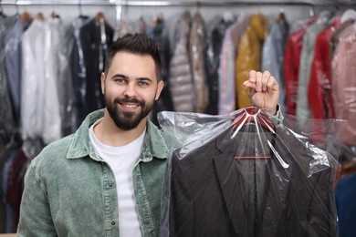 Photo of Dry-cleaning service. Happy man holding hanger with jacket in plastic bag indoors