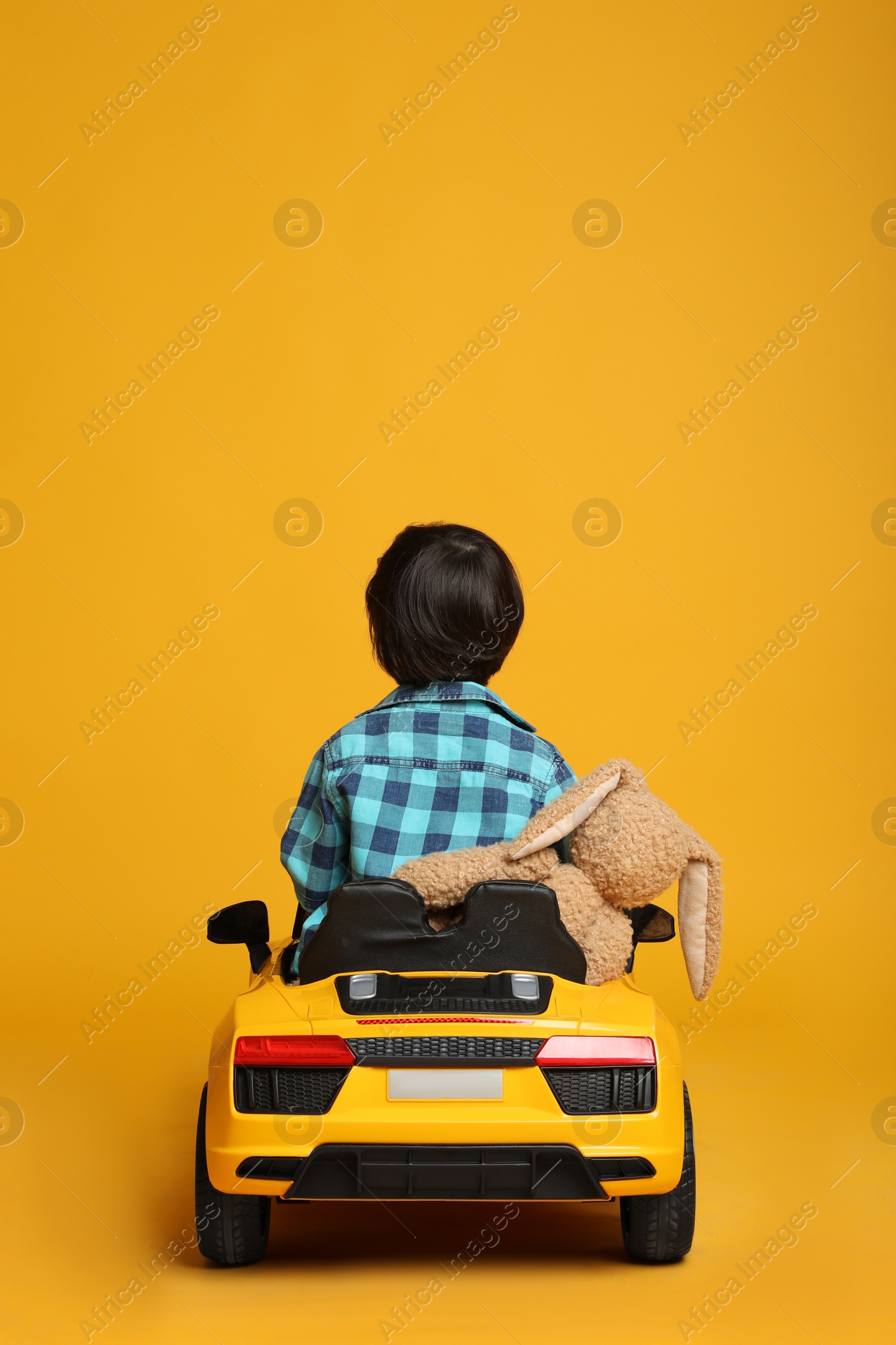 Photo of Cute little boy with toy bunny driving children's car on yellow background, back view