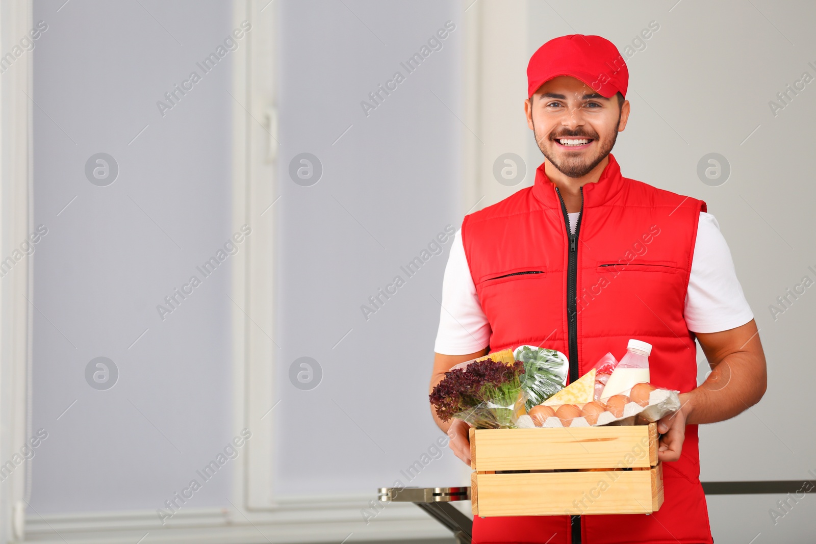 Photo of Food delivery courier holding wooden crate with products indoors. Space for text