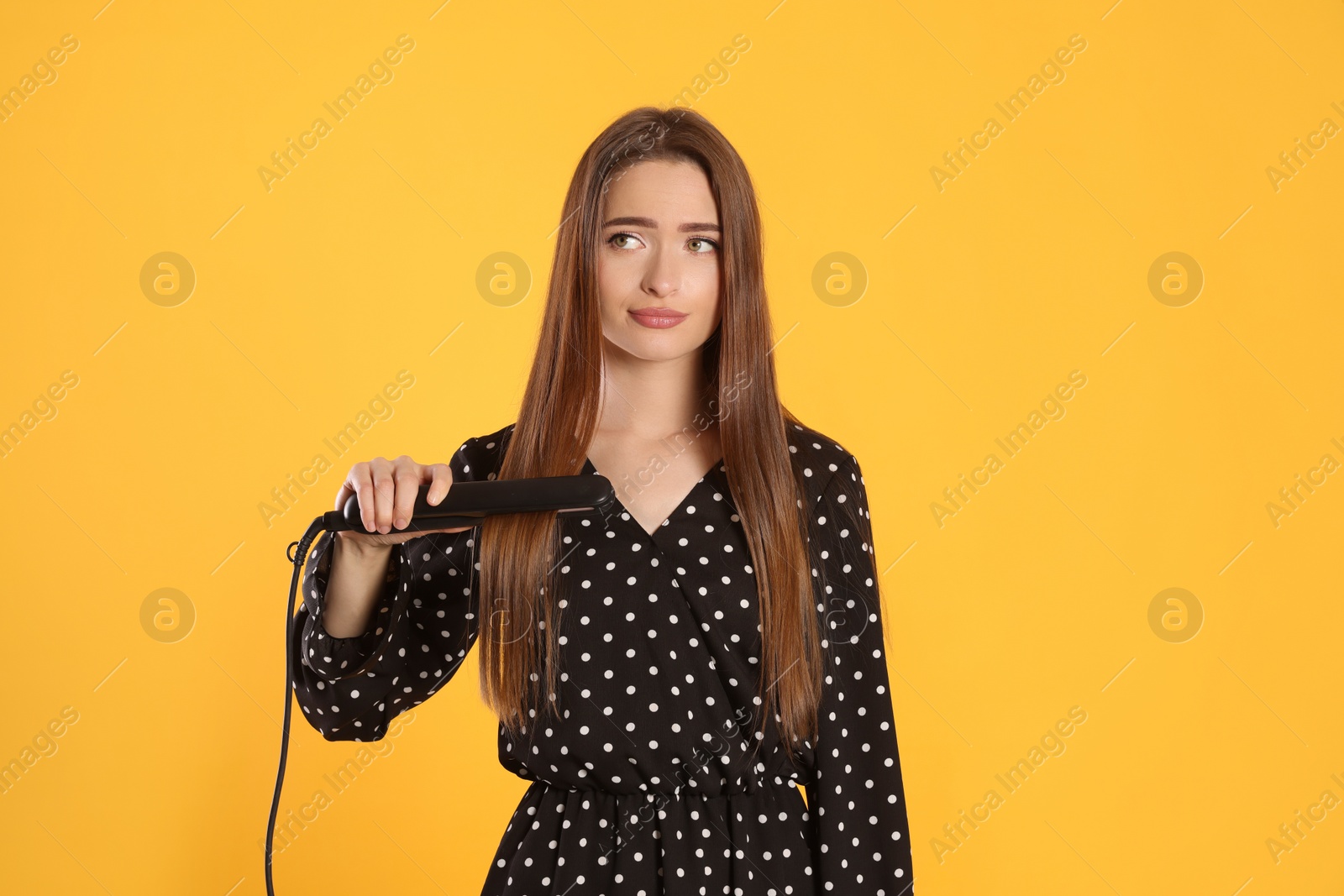 Photo of Upset young woman with flattening iron on yellow background. Hair damage