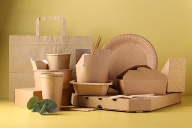 Photo of Eco friendly food packaging. Paper containers, tableware, bag and eucalyptus leaves on pale yellow background