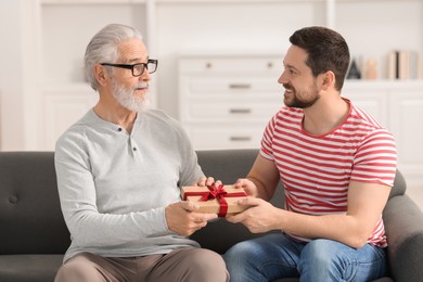 Photo of Son giving gift box to his dad on sofa at home