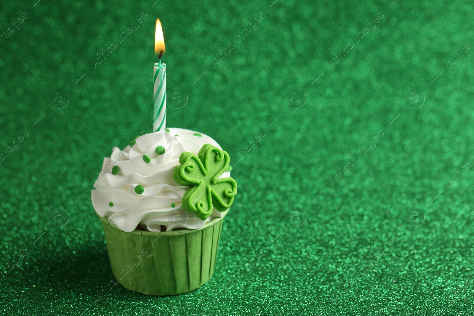 Photo of St. Patrick's day party. Tasty cupcake with clover leaf topper and burning candle on shiny green background. Space for text