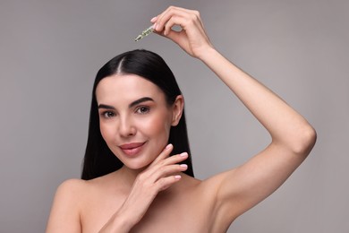 Beautiful young woman using ampoule for hair treatment on grey background