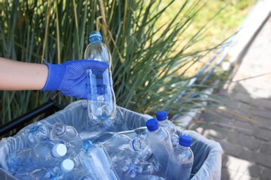 Woman in gloves putting used plastic bottle into trash bin outdoors, closeup. Recycling problem