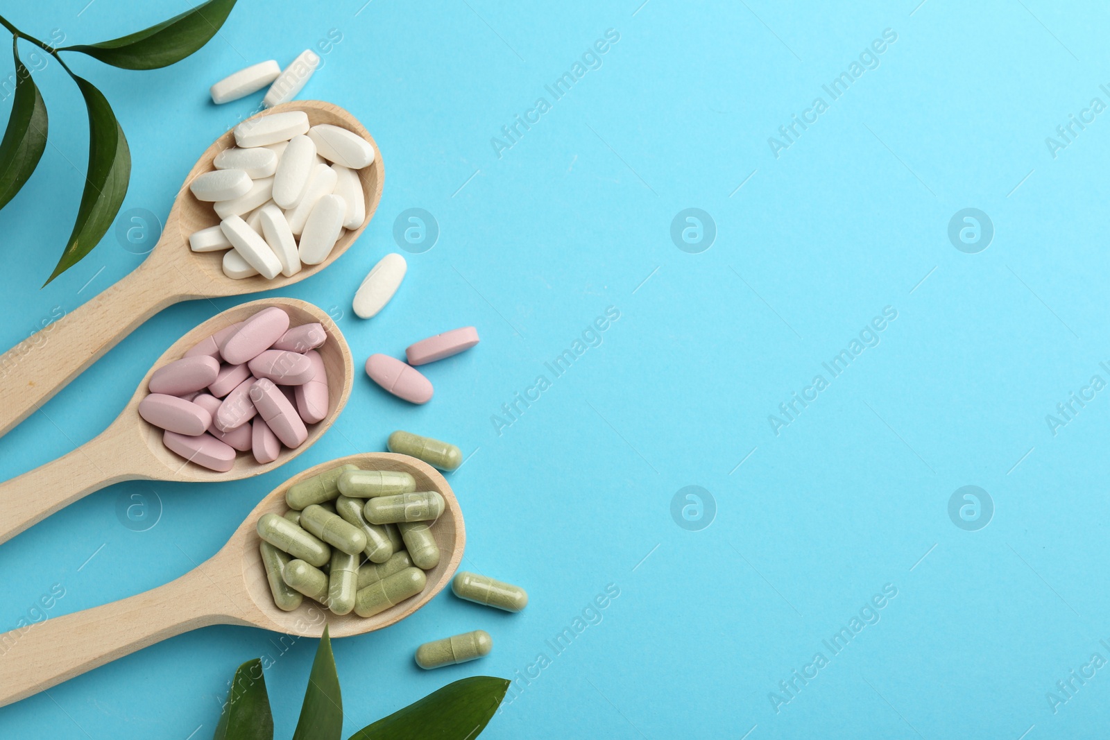 Photo of Different vitamin pills in spoons and green leaves on light blue background, flat lay. Space for text