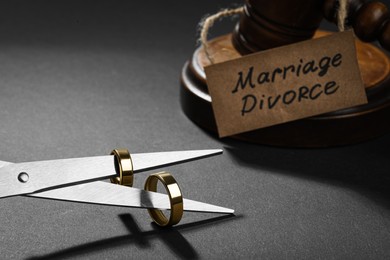 Photo of Scissors with wedding rings on blades near gavel and paper card Marriage Divorce against black background, closeup